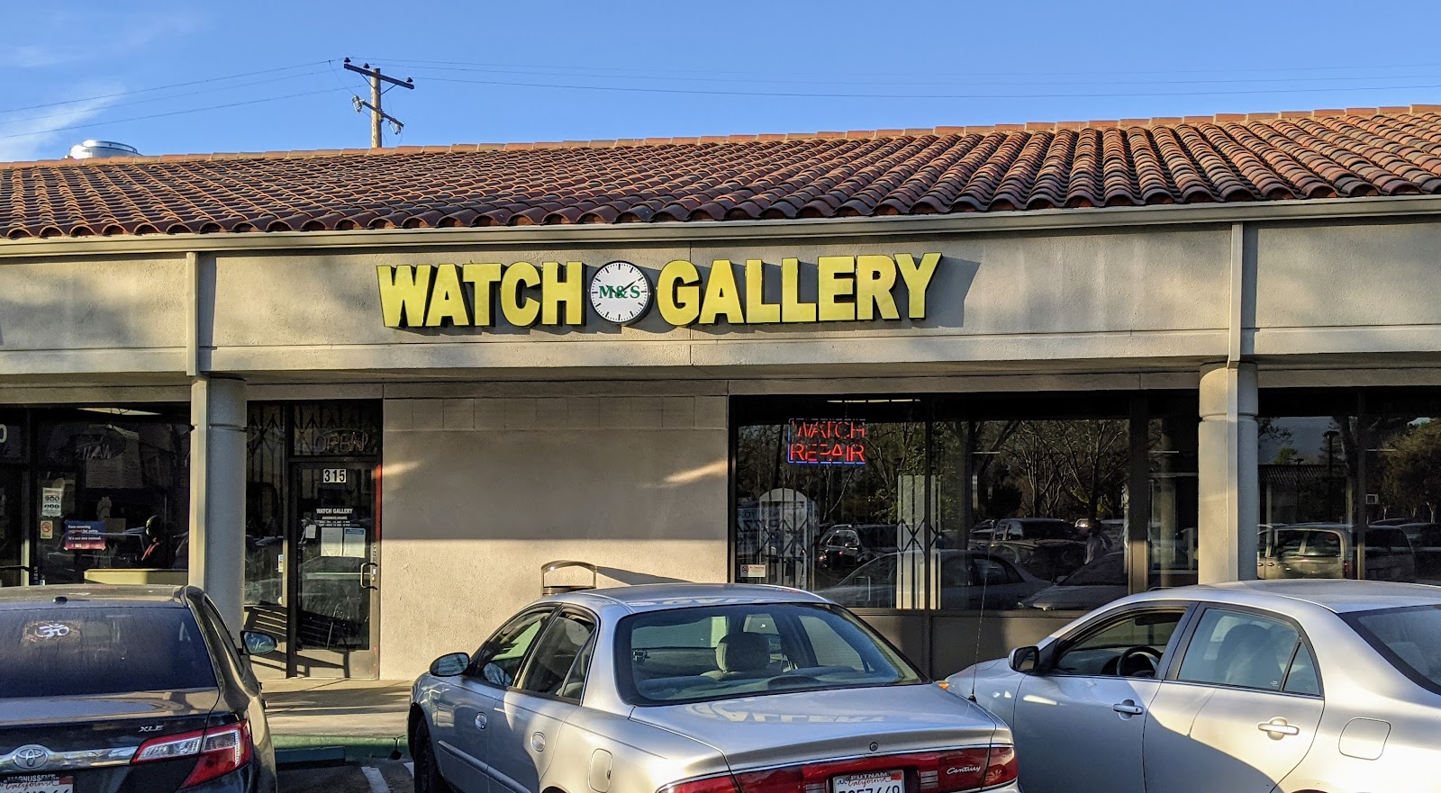M & S Watch Gallery storefront, with the yellow 'WATCH GALLERY' sign on a black backdrop, in Grant Road Plaza, Mountain View.